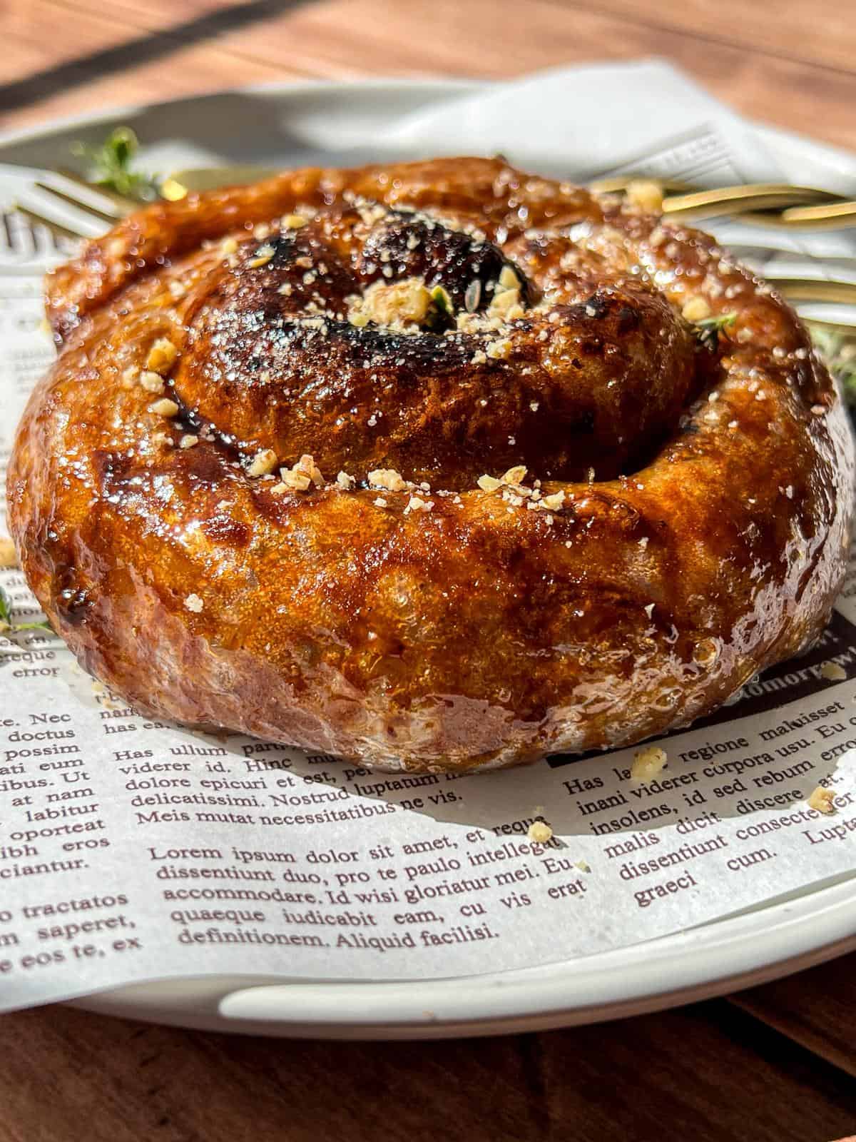 A close-up of a glossy, golden-brown cinnamon roll sprinkled with crushed nuts, served on a plate lined with newspaper-style parchment and two gold forks resting in the background.