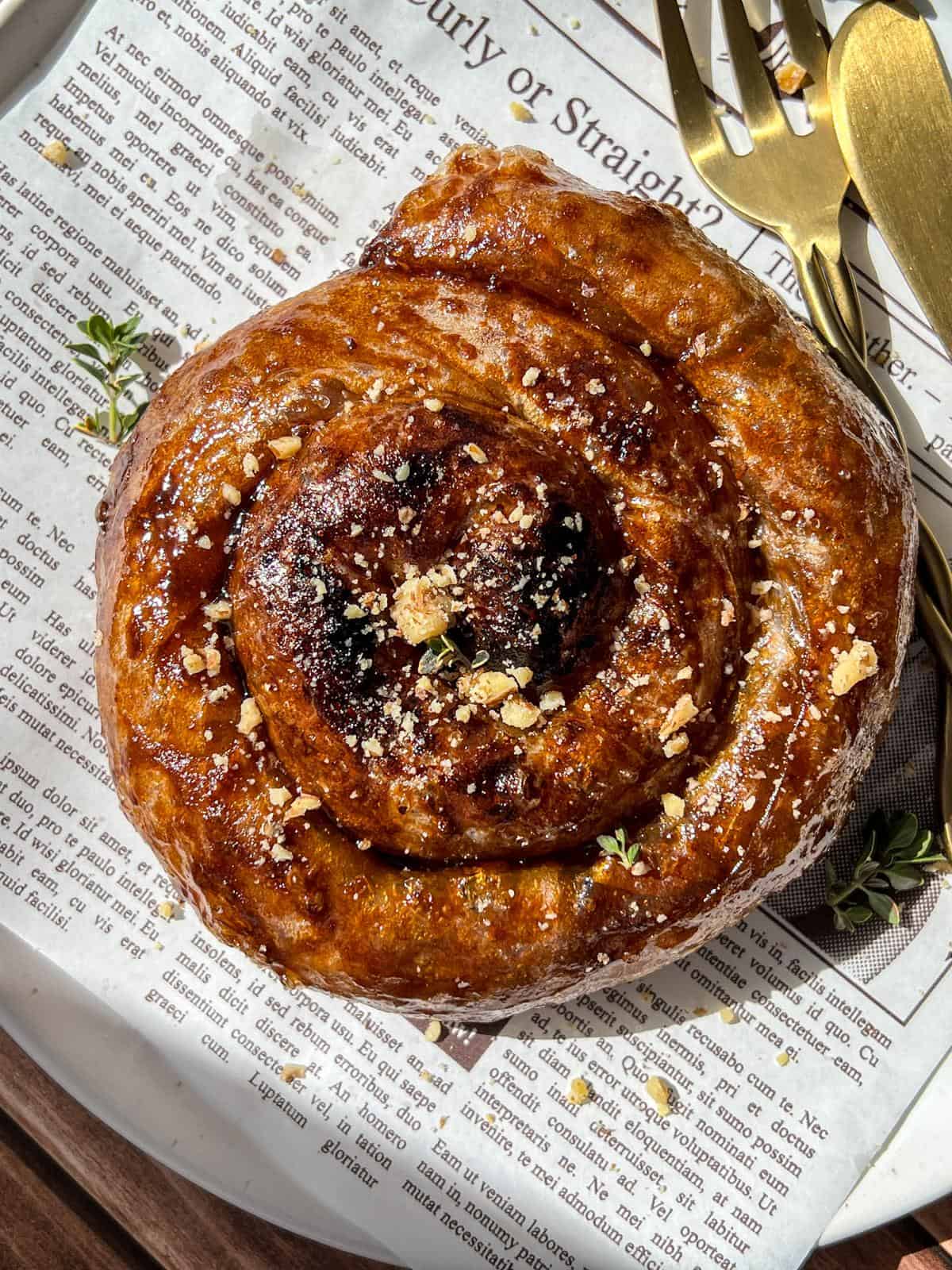 A golden-brown pastry roll topped with crumbs and herbs sits on a plate lined with newspaper print, next to a gold fork and knife. Sunlight casts soft shadows over the scene.
