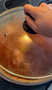 A hand holding the lid of a pot on a stovetop, with condensation visible on the glass lid and some food residue around the pot’s rim.