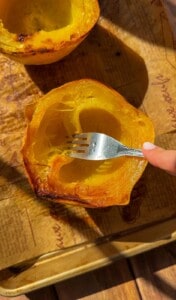 A hand uses a fork to scrape the inside of a roasted spaghetti squash half on a parchment-lined baking sheet.