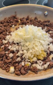 Ground beef is browning in a skillet with a pile of freshly chopped onions and minced garlic on top, ready to be mixed together and cooked further.