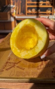A hand holds half of a yellow melon with its seeds removed, revealing the hollow inside. Sunlight highlights the fruit’s juicy texture, and shelves with bread are visible in the background.