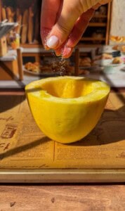 A hand sprinkles salt onto a halved spaghetti squash resting on a baking sheet, with a blurred background of a bakery display and bread.