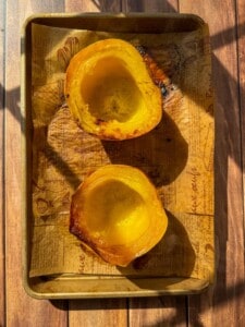 Two roasted acorn squash halves placed cut side up on a baking sheet lined with printed parchment paper, sitting on a wooden surface with sunlight casting shadows.