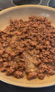 Ground beef browning in a frying pan on a stovetop, with some pieces still slightly pink and juices visible in the pan.