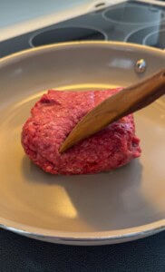 A wooden spatula presses down on a chunk of raw ground beef in a beige frying pan on a stovetop.
