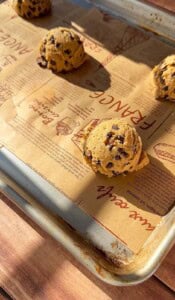 Three scoops of Brown Butter Chocolate Chip Cookie dough sit on a parchment-lined baking tray, ready for baking. Sunlight casts warm shadows across the tray, illuminating the decorative French writing and illustrations on the parchment.