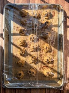 A baking tray lined with parchment paper holds 18 scoops of Brown Butter Chocolate Chip Cookie dough, spaced in rows. The tray, partially covered with plastic wrap, rests on a wooden surface bathed in sunlight.