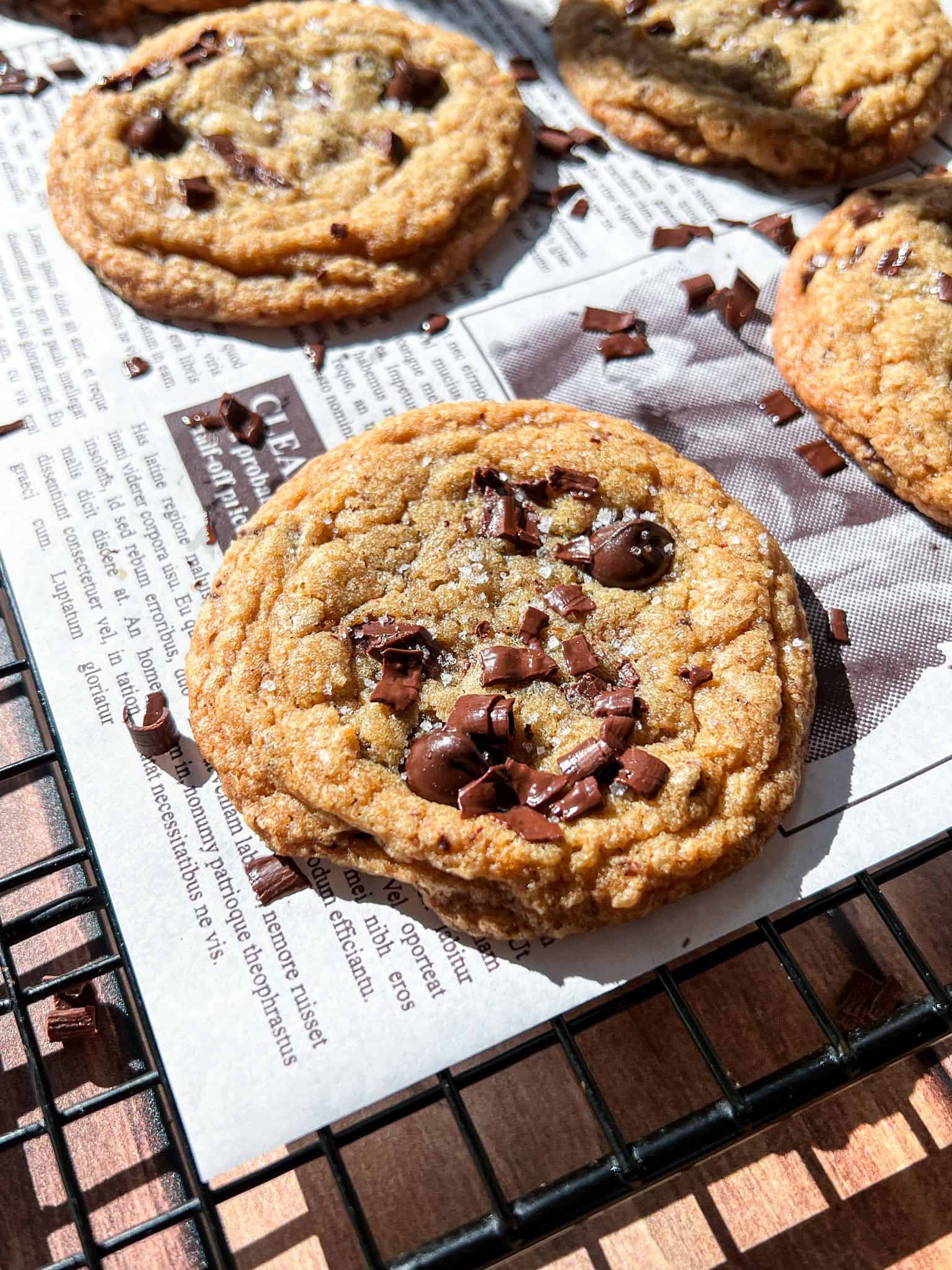 A close-up of a Brown Butter Chocolate Chip Cookie with chocolate chunks on top, resting on printed parchment paper atop a wire cooling rack. Sunlight highlights the cookie’s golden-brown texture.