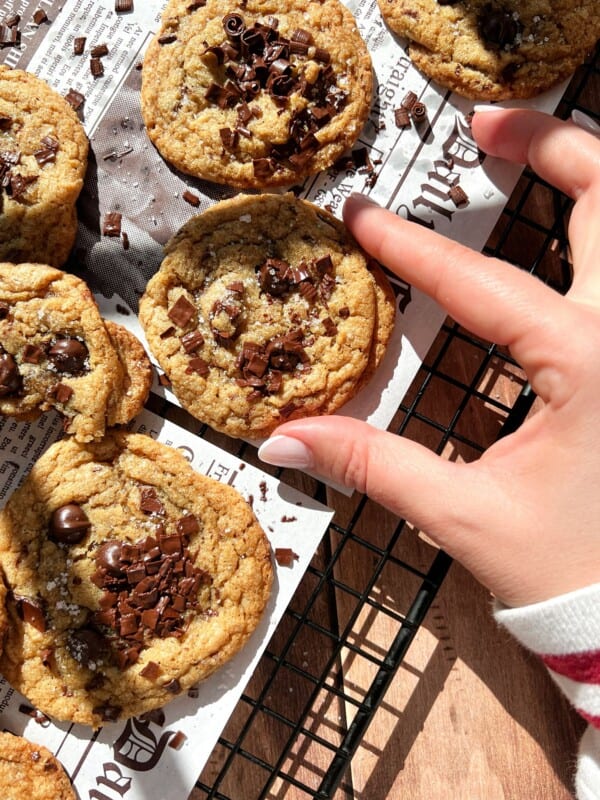 A hand reaches for a Brown Butter Chocolate Chip Cookie on a cooling rack lined with newspaper-style parchment, surrounded by other cookies crowned with chocolate chunks as sunlight casts inviting shadows across the scene.