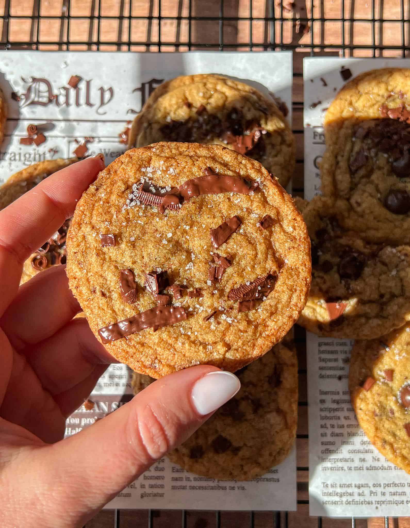 A hand with white-painted nails holds a Brown Butter Chocolate Chip Cookie topped with melted chocolate and sea salt. More cookies are visible on a newspaper-lined surface in the background.