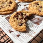 A close-up of Brown Butter Chocolate Chip Cookies on a cooling rack lined with newspaper; one cookie has a large bite taken out, revealing gooey melted chocolate chips inside.
