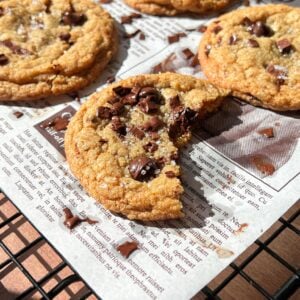 A close-up of Brown Butter Chocolate Chip Cookies on a cooling rack lined with newspaper; one cookie has a large bite taken out, revealing gooey melted chocolate chips inside.