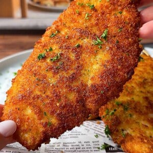 A person holds a large, crispy, golden-brown breaded cutlet garnished with chopped parsley above a plate lined with printed paper.