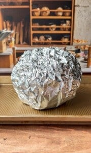 A round object completely wrapped in crinkled aluminum foil sits on a baking sheet with a bakery display of bread loaves in the background.