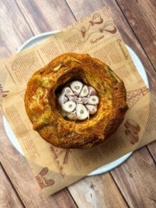A round loaf of bread with a hollow center filled with halved garlic bulbs, placed on brown parchment paper atop a white plate on a wooden surface.