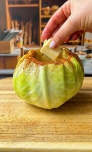 A hand places a pat of butter into a hollowed-out green cabbage sprinkled with spices, set on a wooden cutting board with a kitchen scene blurred in the background.