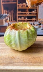A whole green cabbage with the top cut off sits on a wooden surface. Seasoning is being sprinkled onto the open top, and there are shelves with bread and kitchen items blurred in the background.
