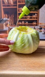 A hand holds a whole cabbage on a wooden cutting board while olive oil is poured over it from a green bottle; shelves with bread are visible in the blurred background.