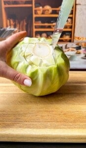 A hand holds a green cabbage on a wooden cutting board while a knife is inserted into the stem, preparing to cut it. Shelves with kitchen items are visible in the background.