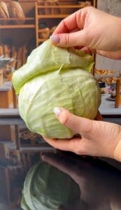 Two hands are peeling an outer leaf from a large, fresh green cabbage with a bakery background blurred in the distance.