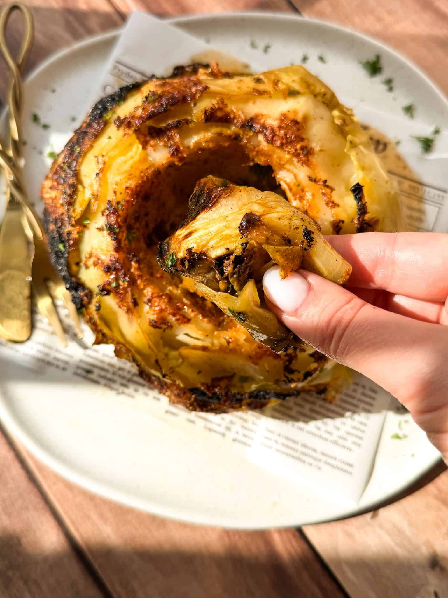A hand holds a roasted cabbage leaf above a plate with a whole, charred roasted cabbage, garnished with herbs. A fork and knife rest on the plate, and sunlight highlights the food.