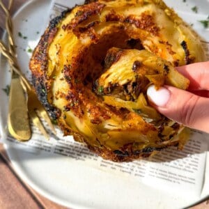 A hand pulls a crispy roasted slice from a whole roasted cabbage served on a white plate with golden cutlery and a piece of printed paper underneath. Sunlight highlights the charred edges and seasoning.