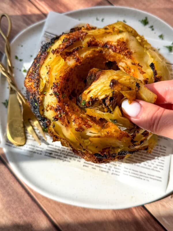 A hand pulls a crispy roasted slice from a whole roasted cabbage served on a white plate with golden cutlery and a piece of printed paper underneath. Sunlight highlights the charred edges and seasoning.