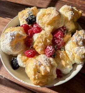 A bowl of golden, fluffy cream puffs dusted with powdered sugar, garnished with fresh raspberries, blackberries, and sprigs of thyme, all bathed in warm sunlight on a wooden surface.