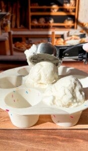 An ice cream scooper is serving two scoops of vanilla ice cream into a white tray with sprinkle designs, with a bakery display blurred in the background.