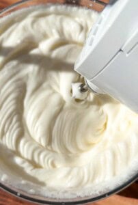 A close-up of an electric hand mixer whipping a creamy, white mixture in a glass bowl, creating smooth, fluffy peaks.