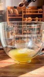 A thin stream of liquid is pouring from an espresso machine into a clear KitchenAid mixing bowl containing a yellow mixture, with shelves of bread in the background.