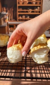 A hand with light-pink nails lifts a fluffy, golden-topped baked meringue from a cooling rack, with more meringues and shelves of bread in the blurred background.