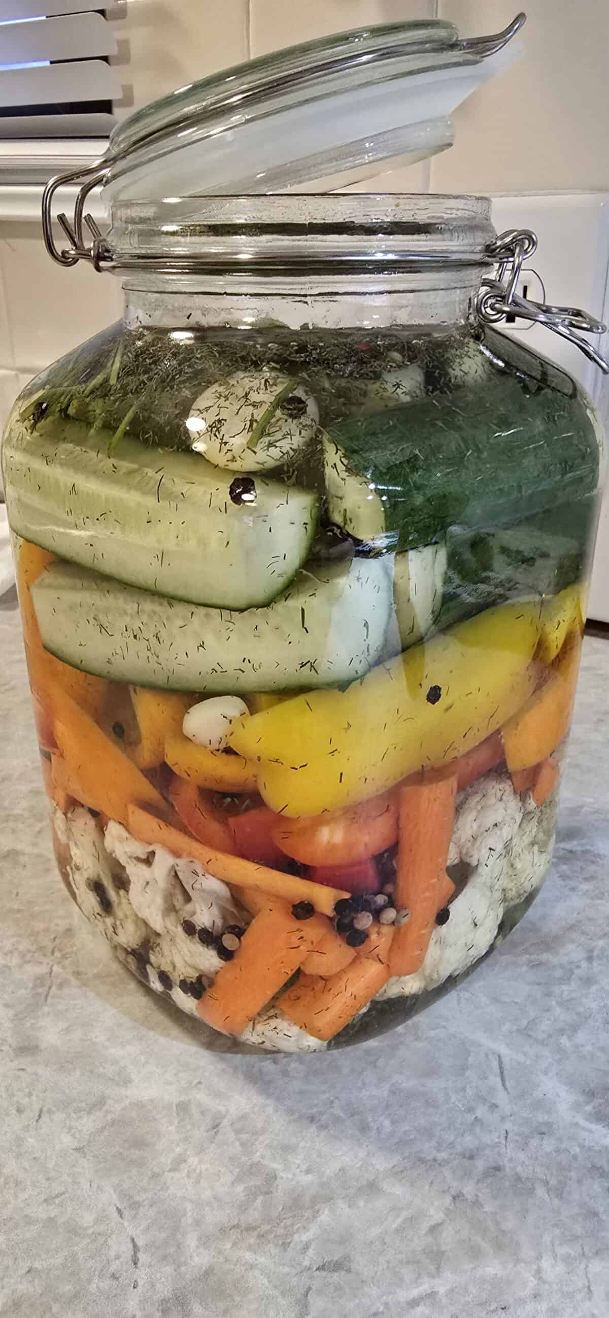 A large glass jar filled with quick pickled veggies—cucumbers, carrots, cauliflower, and yellow peppers—sits on a kitchen countertop. The jar has a hinged lid and is filled with brine and herbs.
