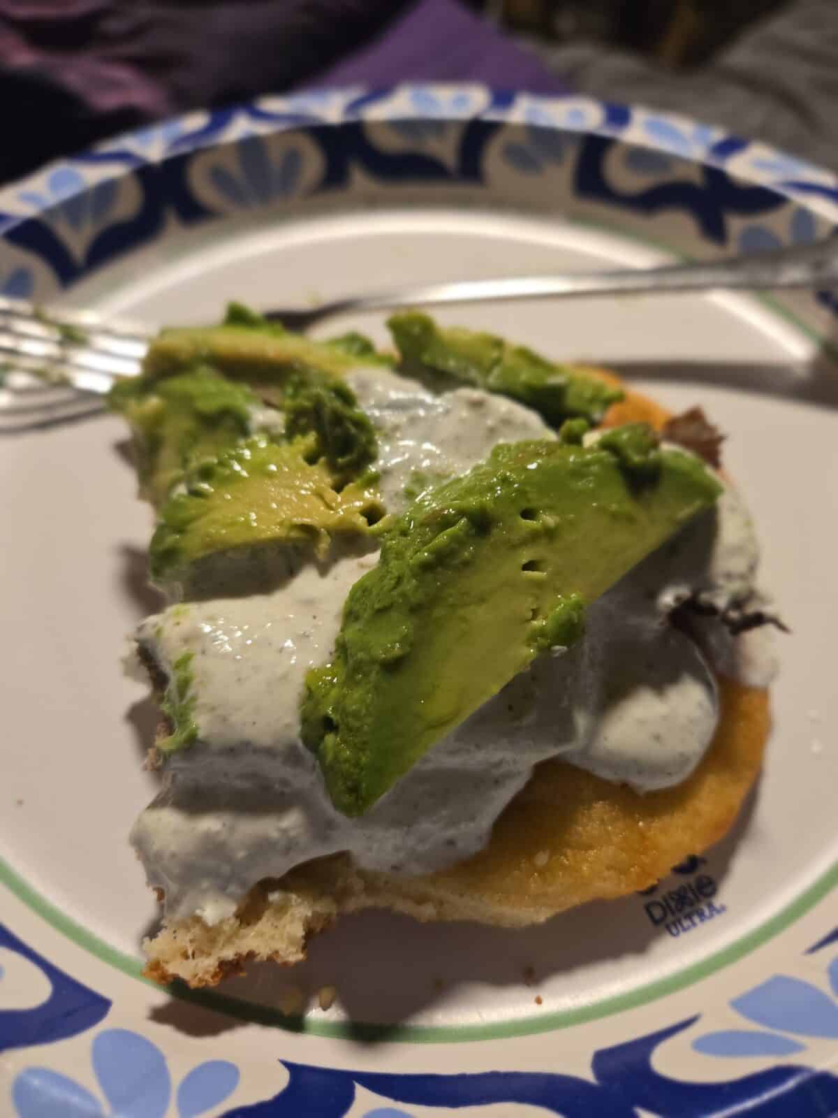 A paper plate holds a piece of bread topped with avocado slices and a creamy white sauce, inspired by the viral HOMEMADE DONER KEBAB trend. A fork rests on the plate beside the food.