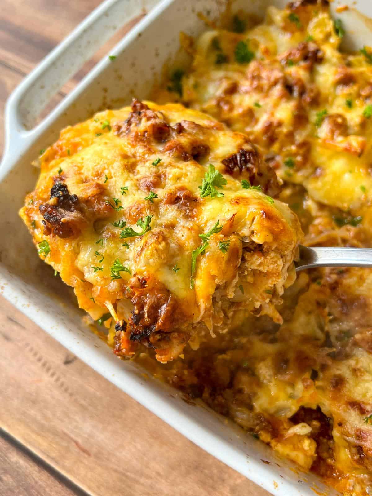 A close-up of a cheesy, golden-brown Cabbage Lasagna being lifted from a baking dish with a spoon, topped with melted cheese and garnished with chopped parsley. - 3
