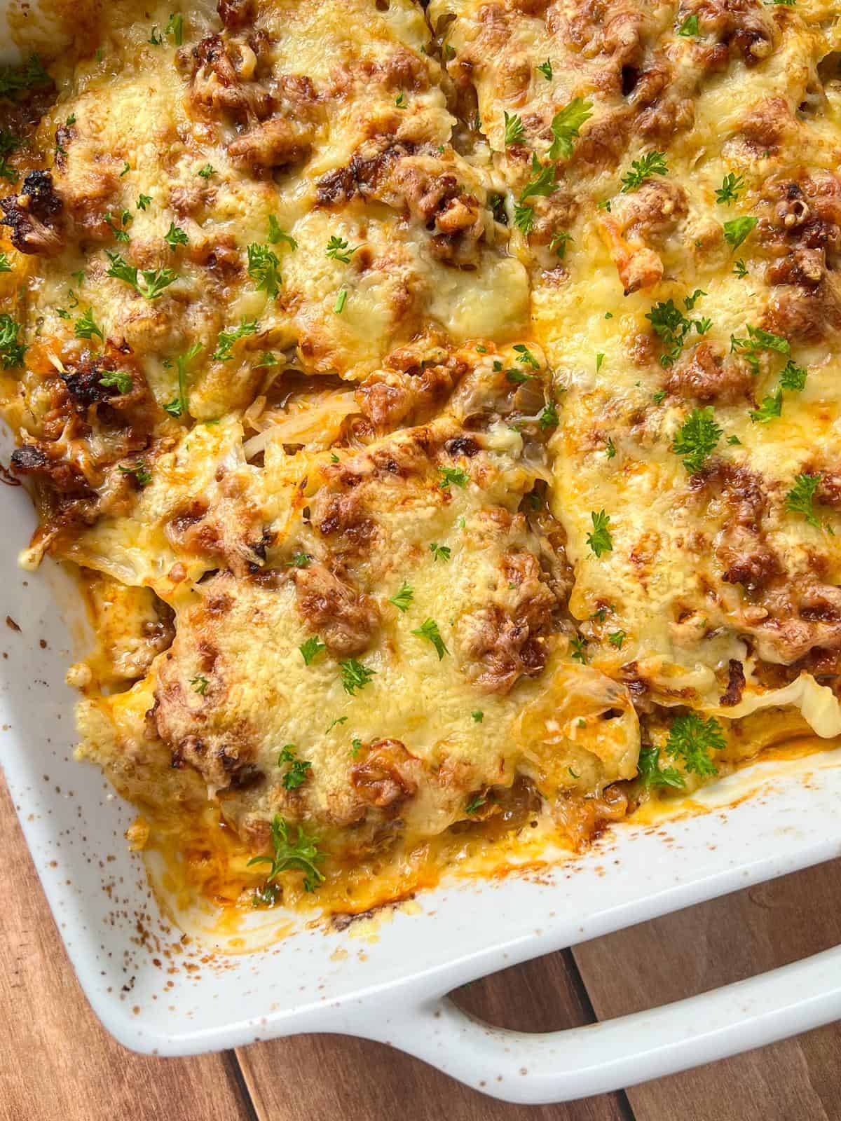 A close-up of a Cabbage Lasagna casserole in a white dish, topped with melted cheese, browned ground meat, and sprinkled with fresh chopped parsley on a wooden surface. - 4