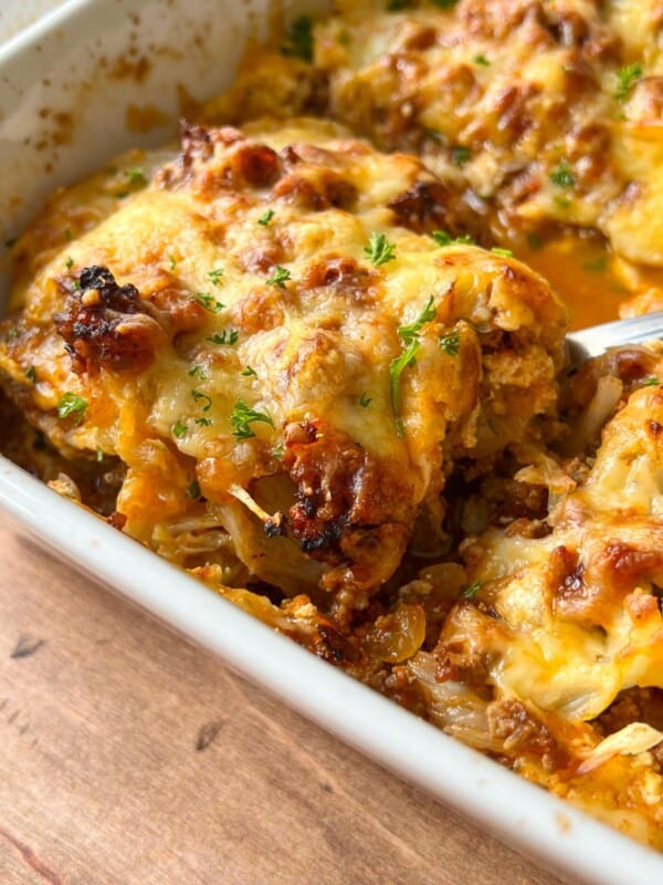 Close-up of a baked Cabbage Lasagna in a white dish, featuring a golden, melted cheese topping with browned edges and sprinkled with fresh chopped herbs. A serving spoon is lifting a portion from the dish.