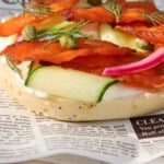 An open-faced bagel topped with cream cheese, smoked salmon jerky, capers, pickled red onions, fresh dill, and thin slices of zucchini, served on a plate lined with newspaper-print parchment.