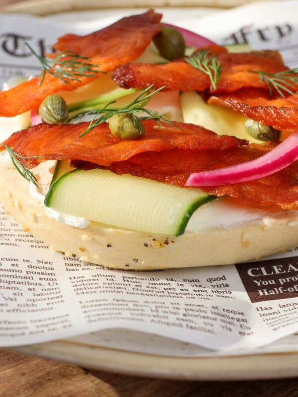 An open-faced bagel topped with cream cheese, smoked salmon jerky, capers, pickled red onions, fresh dill, and thin slices of zucchini, served on a plate lined with newspaper-print parchment.