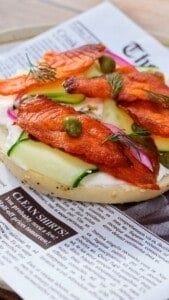 An open-faced bagel topped with cream cheese, smoked salmon jerky, cucumber slices, capers, pickled onions, and fresh dill, served on newspaper-style parchment paper.