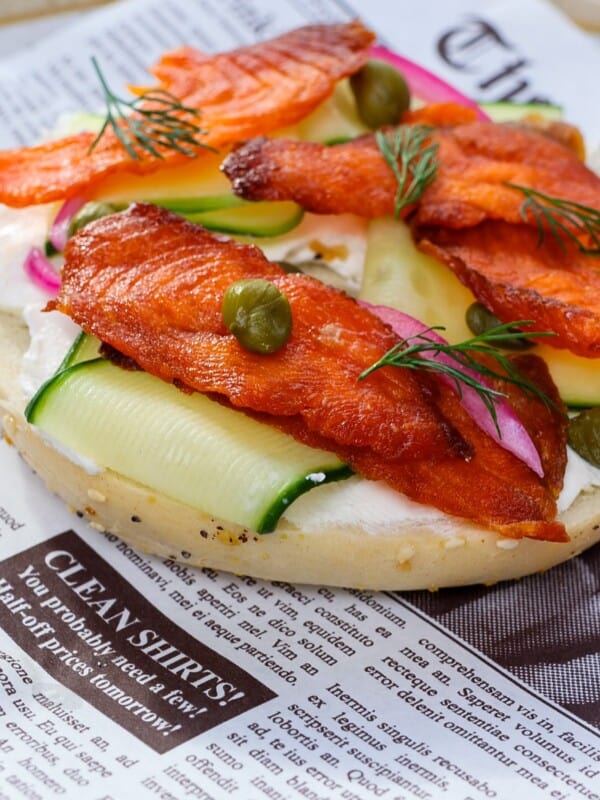 An open-faced bagel topped with cream cheese, smoked salmon jerky, cucumber slices, capers, pickled onions, and fresh dill, served on newspaper-style parchment paper.