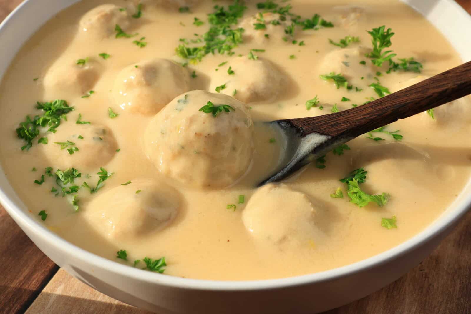 A bowl of meatballs covered in creamy sauce, garnished with chopped parsley. A wooden spoon rests in the bowl, surrounded by the sauce and meatballs. - 3