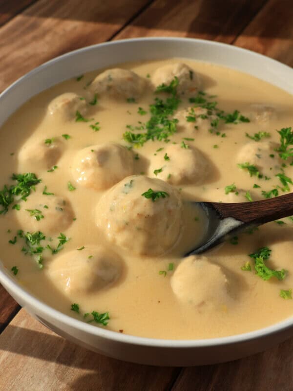 A bowl of creamy sauce with meatballs, garnished with chopped fresh parsley, sits on a wooden table. A wooden spoon is partially submerged in the sauce, ready to serve.