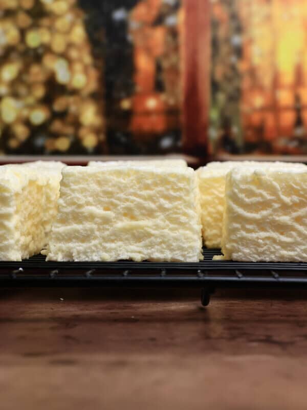 Close-up of fluffy, white rectangular marshmallows arranged on a cooling rack, with a blurred, warm-toned background featuring bokeh lights and wooden elements.