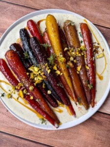 A plate of colorful roasted carrots, topped with chopped pistachios and herbs, served on a creamy white sauce and drizzled with a golden glaze. The plate sits on a wooden surface.