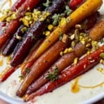 A plate of colorful roasted carrots topped with chopped pistachios and fresh herbs, served on a creamy white sauce, drizzled with oil on a wooden table.