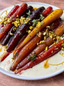 A plate of colorful roasted carrots topped with chopped pistachios and fresh herbs, served on a creamy white sauce, drizzled with oil on a wooden table.