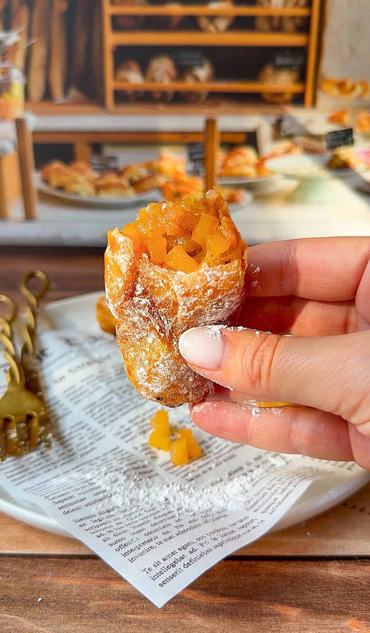A hand holds a Healthy Copycat McDonald's Apple Pie filled with diced orange fruit, dusted with powdered sugar. The pastry sits on a white plate with newspaper print, surrounded by more fruit and sugar, with a bakery display in the background. - 3