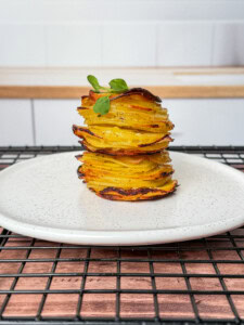 A neatly stacked tower of greek lemon potato stacks, thinly sliced and roasted, garnished with a small green herb, served on a white plate with a speckled pattern, placed on a black cooling rack.